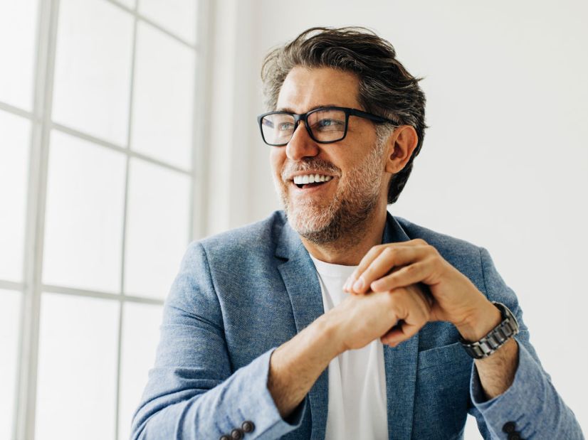 Smiling man in glasses and blue blazer, looking to the side with hands clasped, seated by a bright window.