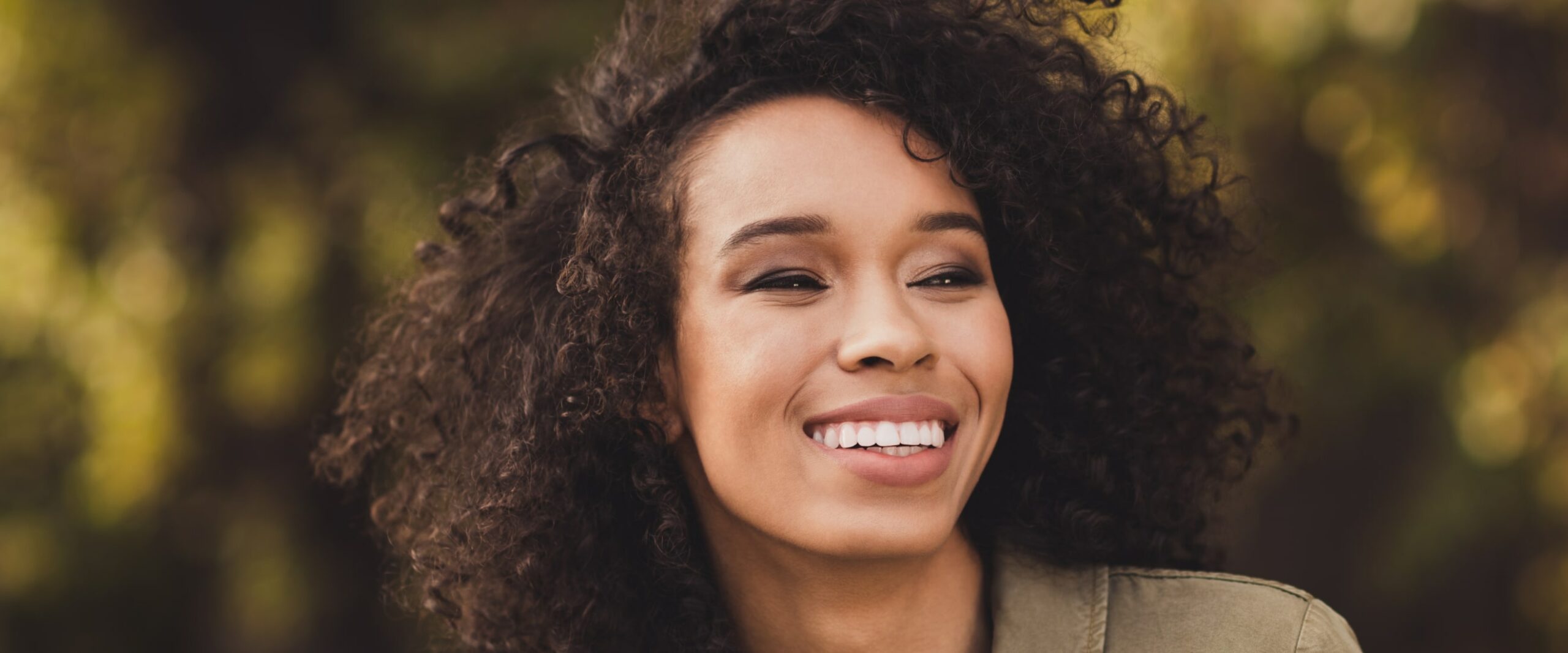 Smiling woman with curly hair looking off-camera, outdoors with a softly blurred background.