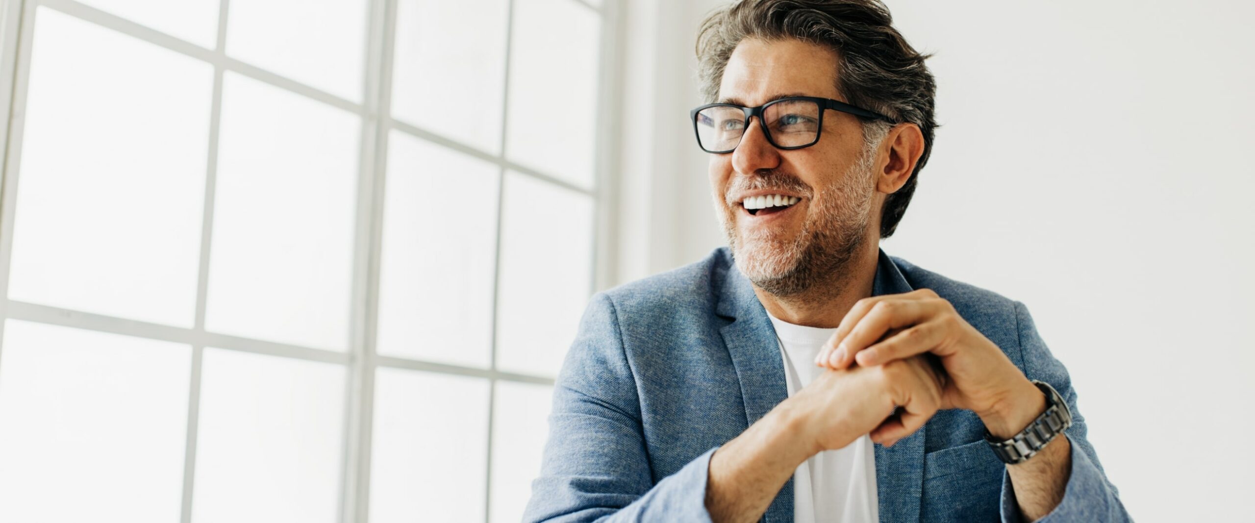 Smiling man with glasses and a beard, wearing a light blue blazer, looking to the side near a large window.