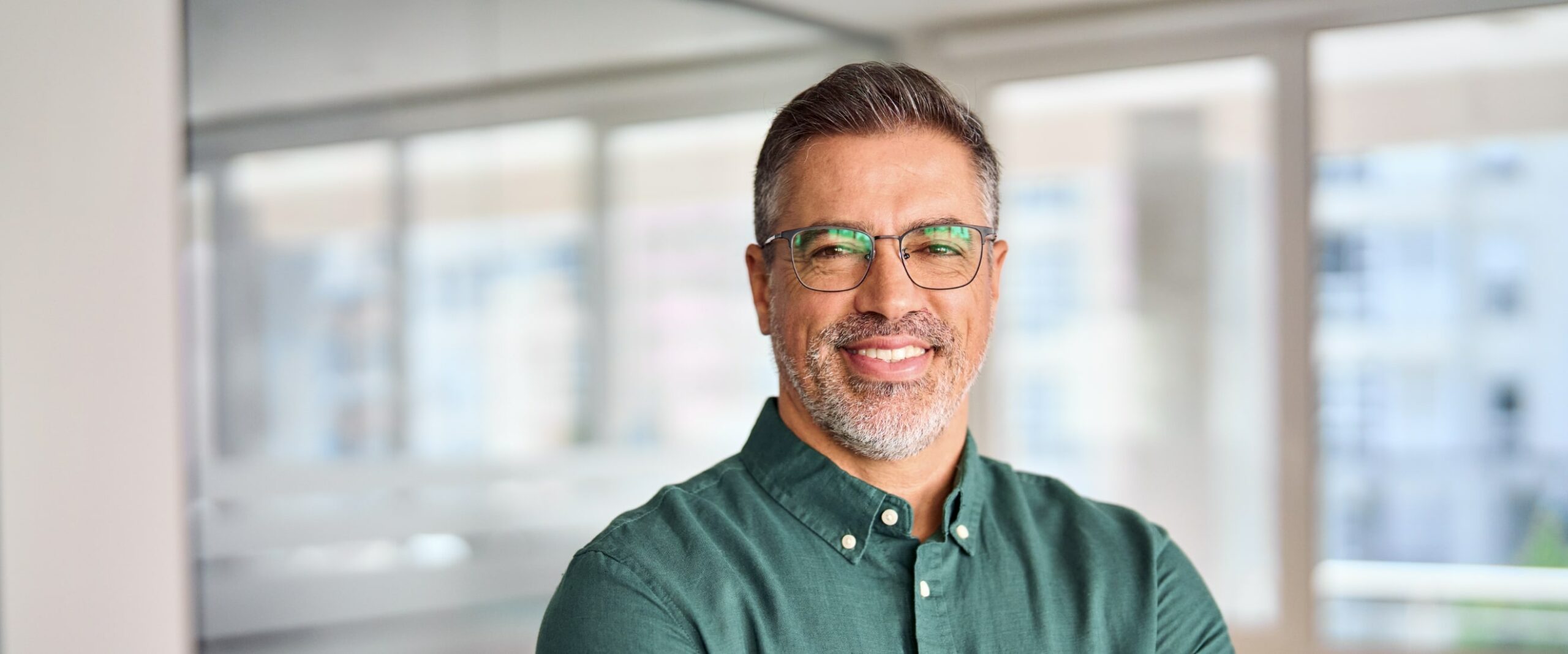 Smiling man with glasses and a beard, wearing a green shirt, standing in a bright office with large windows.