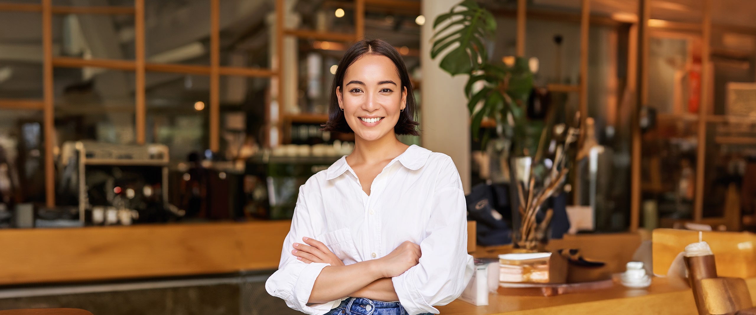 Smiling woman in a white shirt standing with arms crossed in a warmly lit café or workspace.
